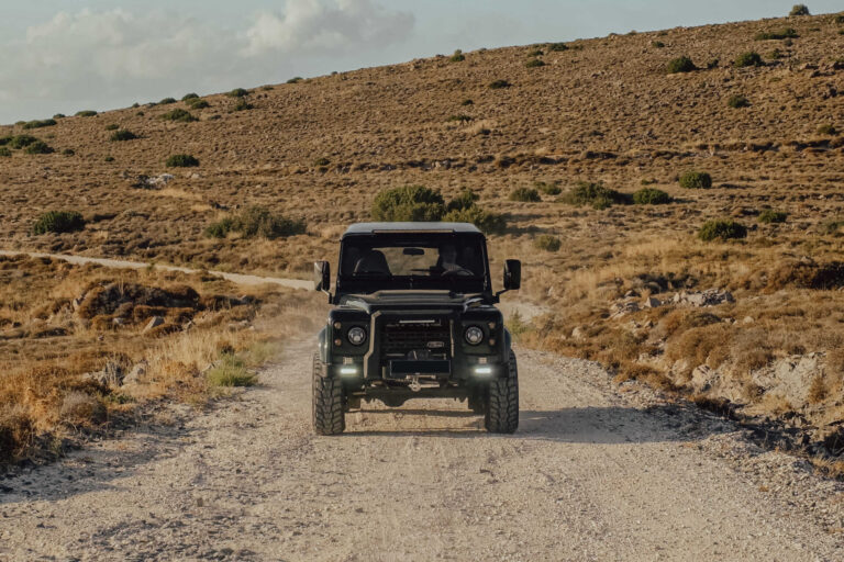 A rugged black Land Rover Defender with off-road modifications drives along a dusty gravel road in an arid, hilly landscape with scattered green shrubs under a partly cloudy sky.