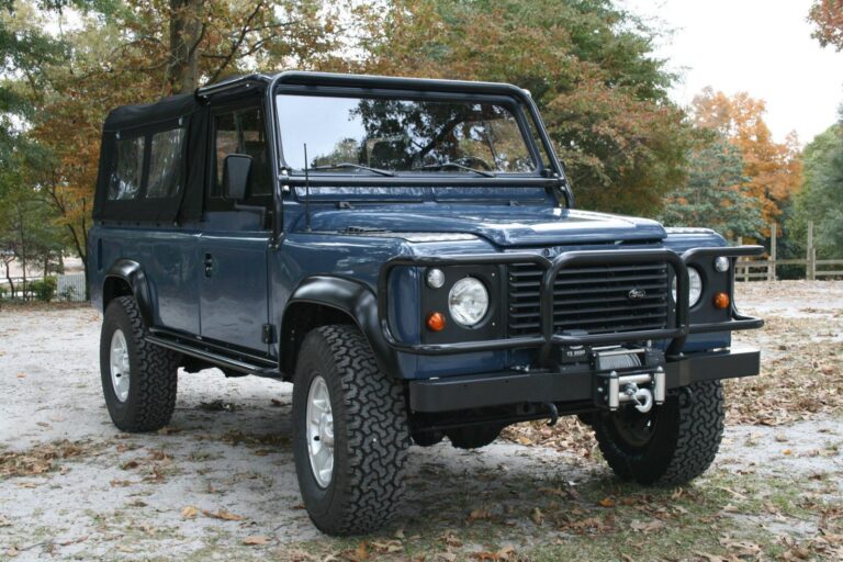 A restored blue Land Rover Defender 110 convertible with a black soft top, rugged off-road tires, and a heavy-duty front bumper with a winch, parked on a gravel surface with autumn foliage in the background.