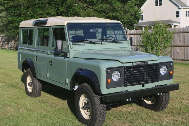 A restored Land Rover Defender 110 in pastel green with a cream roof, parked on a well-maintained grassy lawn. The vehicle features a classic design with black fender flares, rugged off-road tires, and a black front grille. The background includes a wooden fence, green trees, and a suburban house, adding to the vintage yet practical appeal of this iconic 4x4.