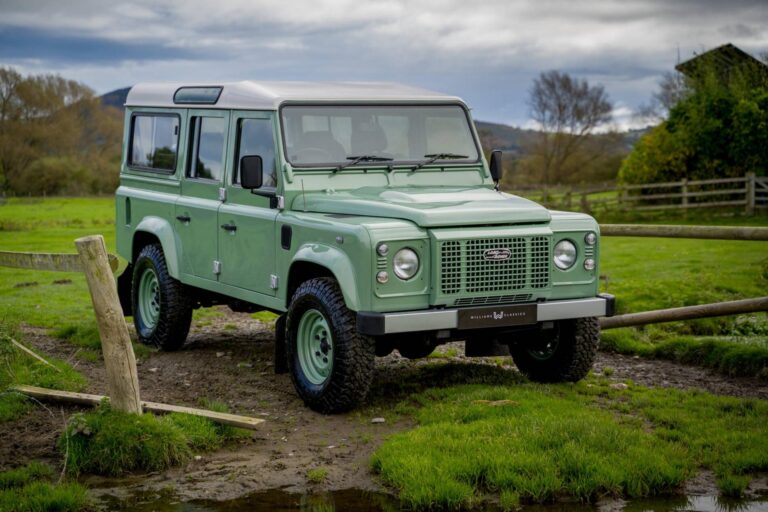 A classic Land Rover Defender 110 in pastel green with a white roof, parked on a muddy countryside trail near a wooden fence. The rugged vehicle, equipped with off-road tires, is set against a scenic rural backdrop with rolling hills, lush green grass, and a cloudy sky.
