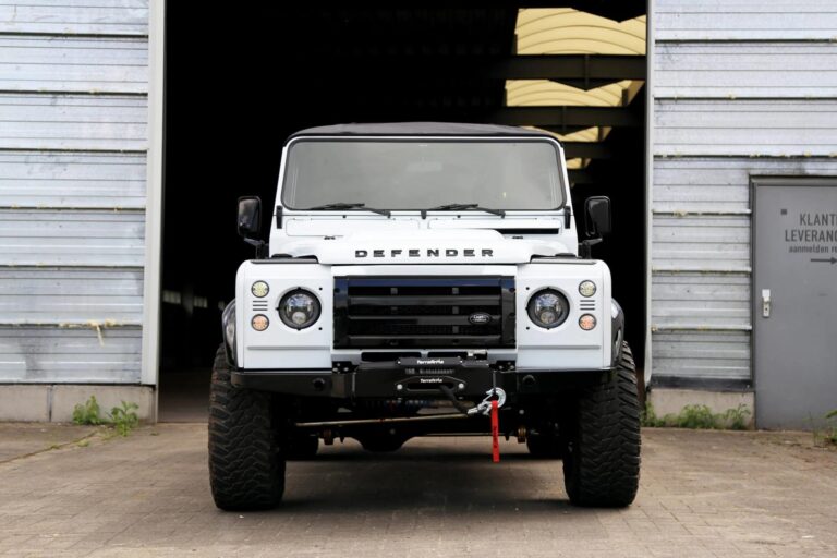 A white Land Rover Defender 110 with a black grille and heavy-duty off-road tires, parked in front of an industrial garage with metallic siding. The vehicle features a front-mounted winch, LED headlights, and a rugged, lifted suspension for enhanced off-road performance. The bold 'DEFENDER' badge is prominently displayed on the hood, emphasizing the classic yet modern appeal of this iconic 4x4.