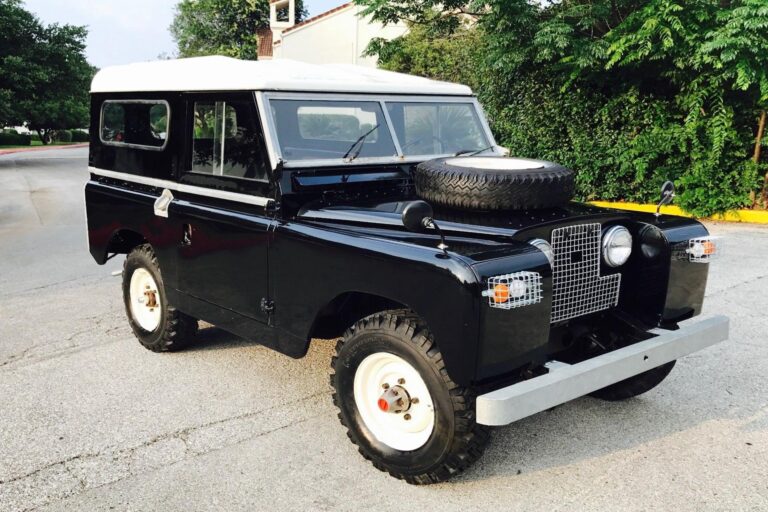 A fully restored 1967 Land Rover Series 2A in glossy black with a white roof, parked on a suburban road. The classic off-roader features a front-mounted spare tire on the hood, white steel wheels with rugged all-terrain tires, and protective metal grilles over the headlights. The vehicle's vintage design, complete with an aluminum front bumper and side mirrors mounted on the fenders, highlights its timeless rugged appeal. Lush green foliage and a residential building can be seen in the background.